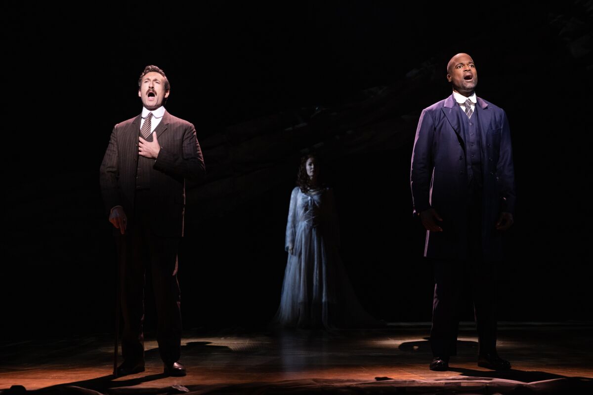 Two actors, spotlighted in the foreground, sing about a figure in half-light behind them.