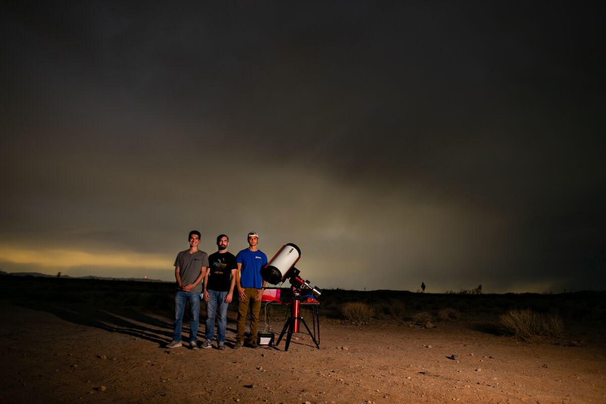 Three people stand in the desert with a telescope