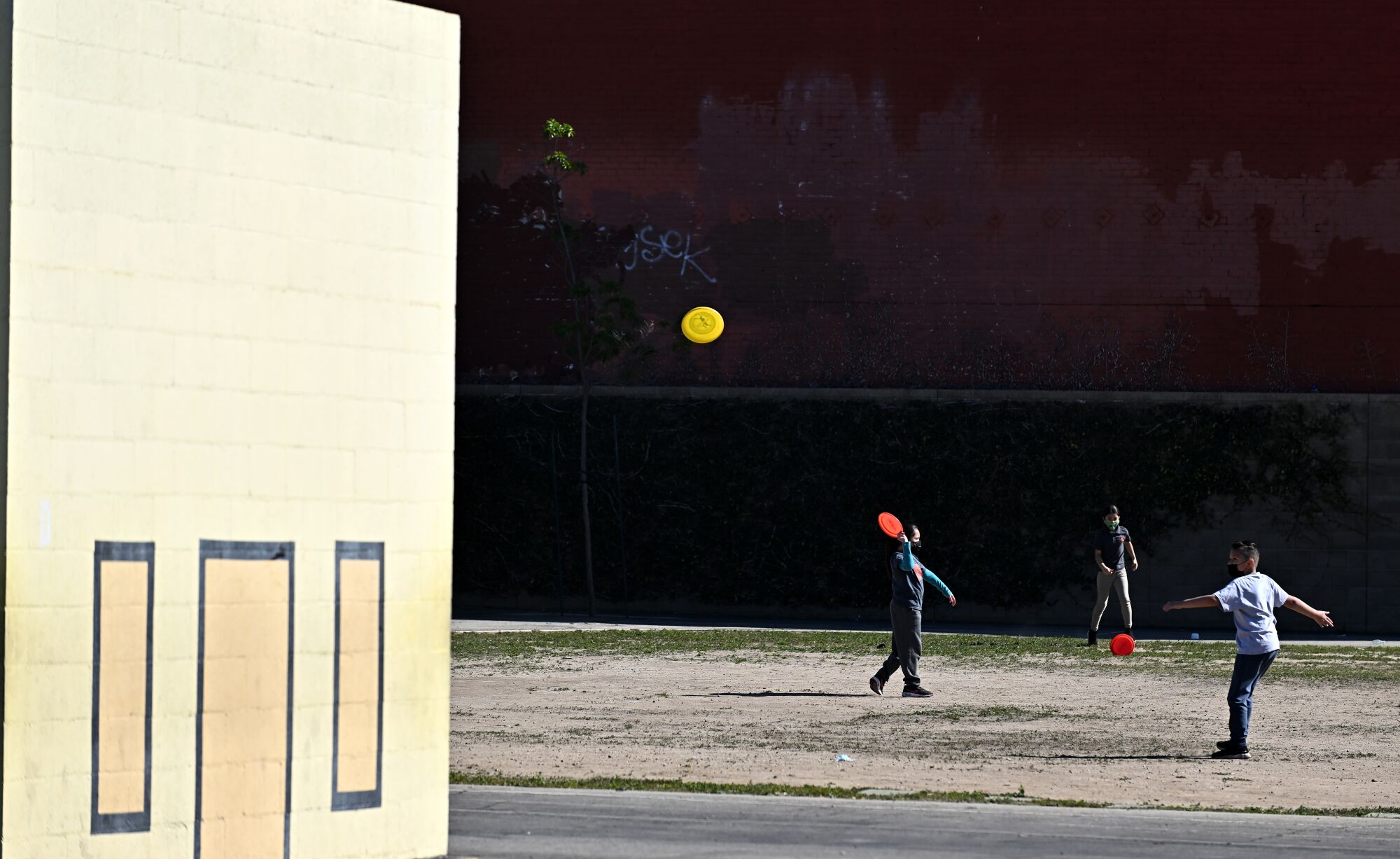 Kids playing with frisbees in a grass-less open field next to a playground wall at a school.