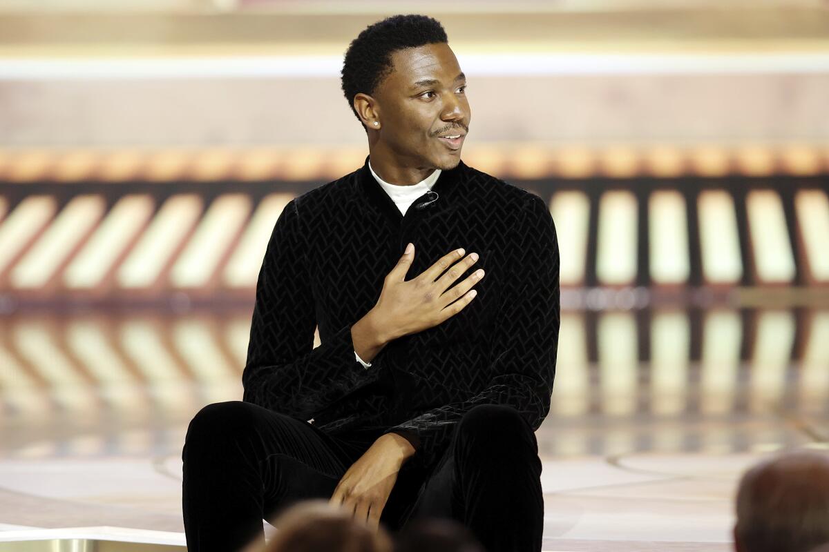 A man in casual-chic attire sits on a stage with his right hand over his heart