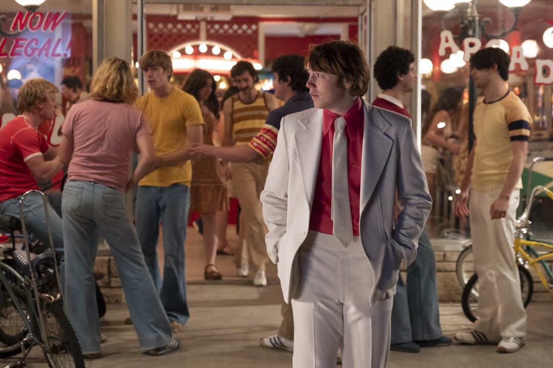 A young man in a white suit and tie hangs out in front of an arcade in "Licorice Pizza.