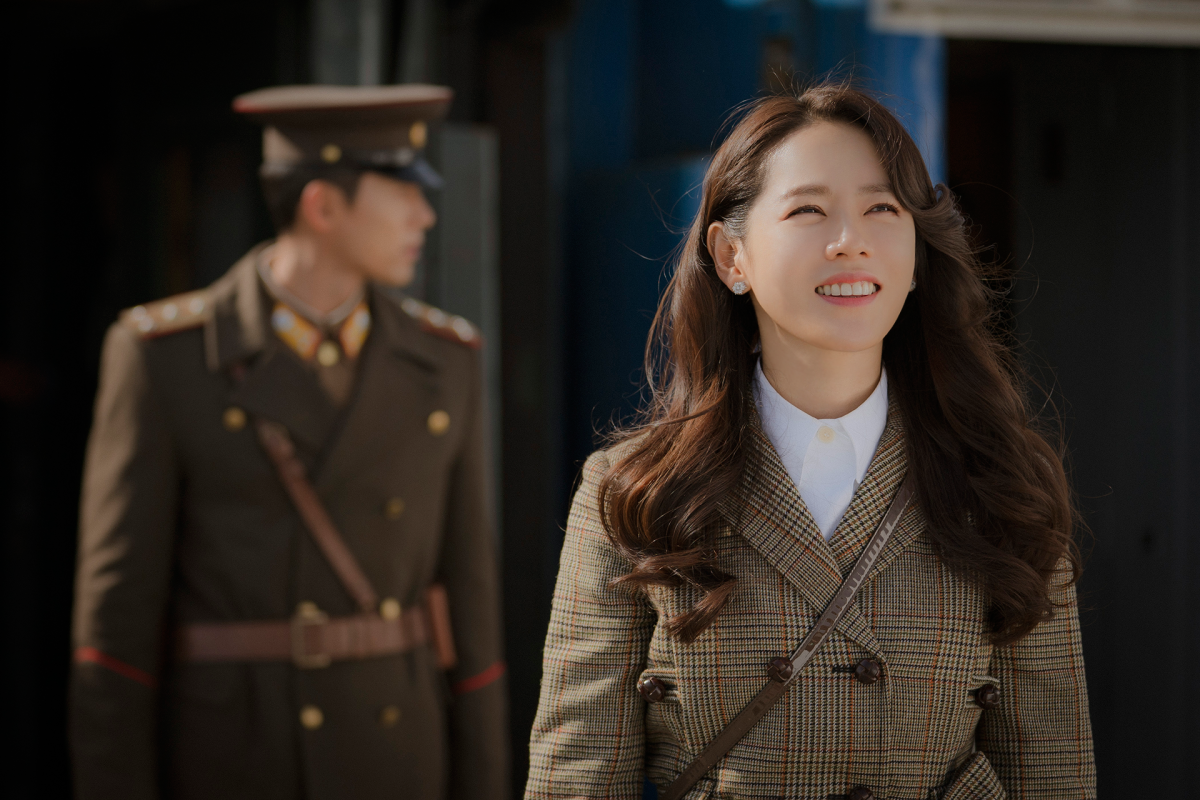 A woman looks up to the sky smiling, with a soldier in the background.