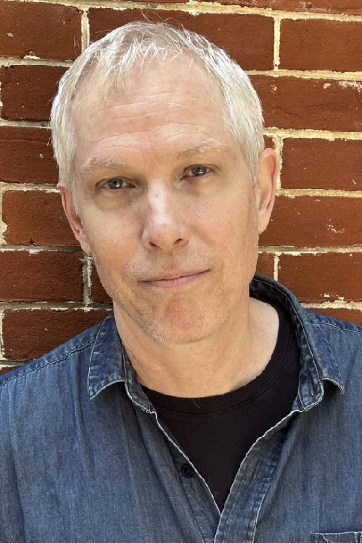 A white man with grey hair in front of a brick wall