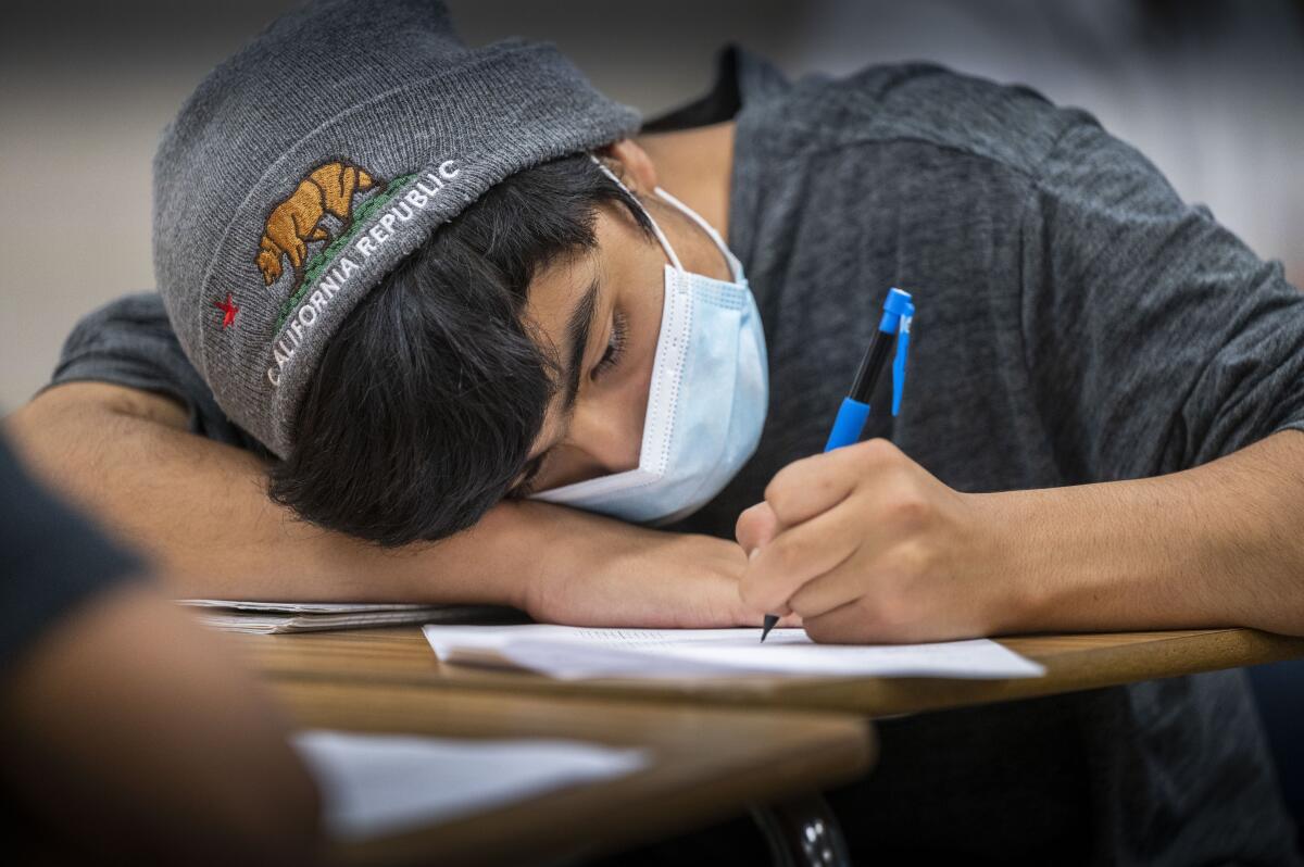 A teenager sitting at a desk lays his head on one arm while writing.