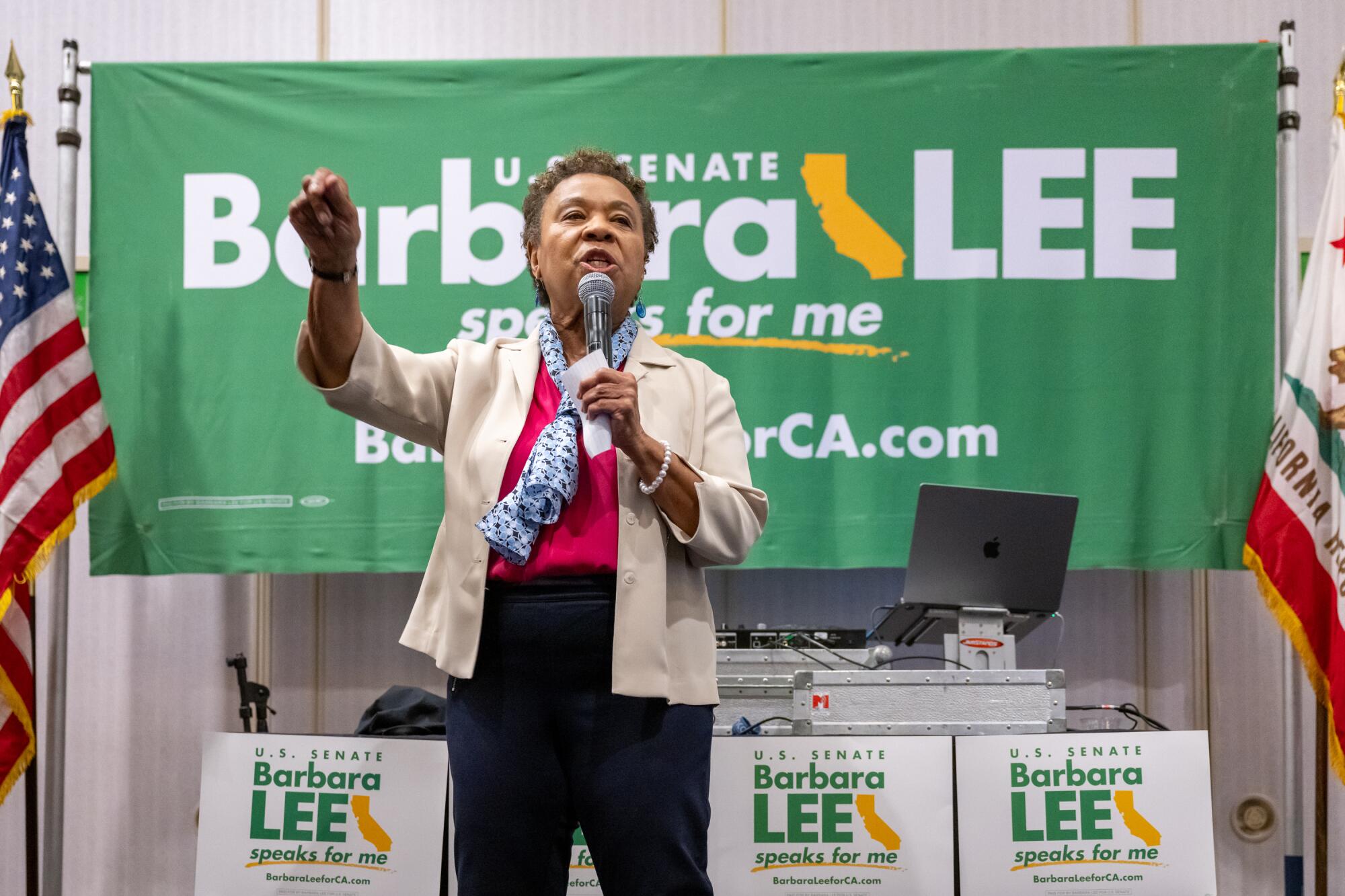 Rep. Barbara Lee gestures as she stands in front of a banner and signs for her Senate campaign, speaking into a microphone.