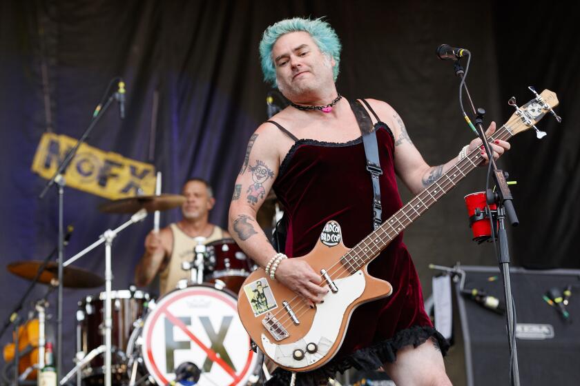 A man with blue hair wearing a dress plays the bass on stage while another man drums behind him