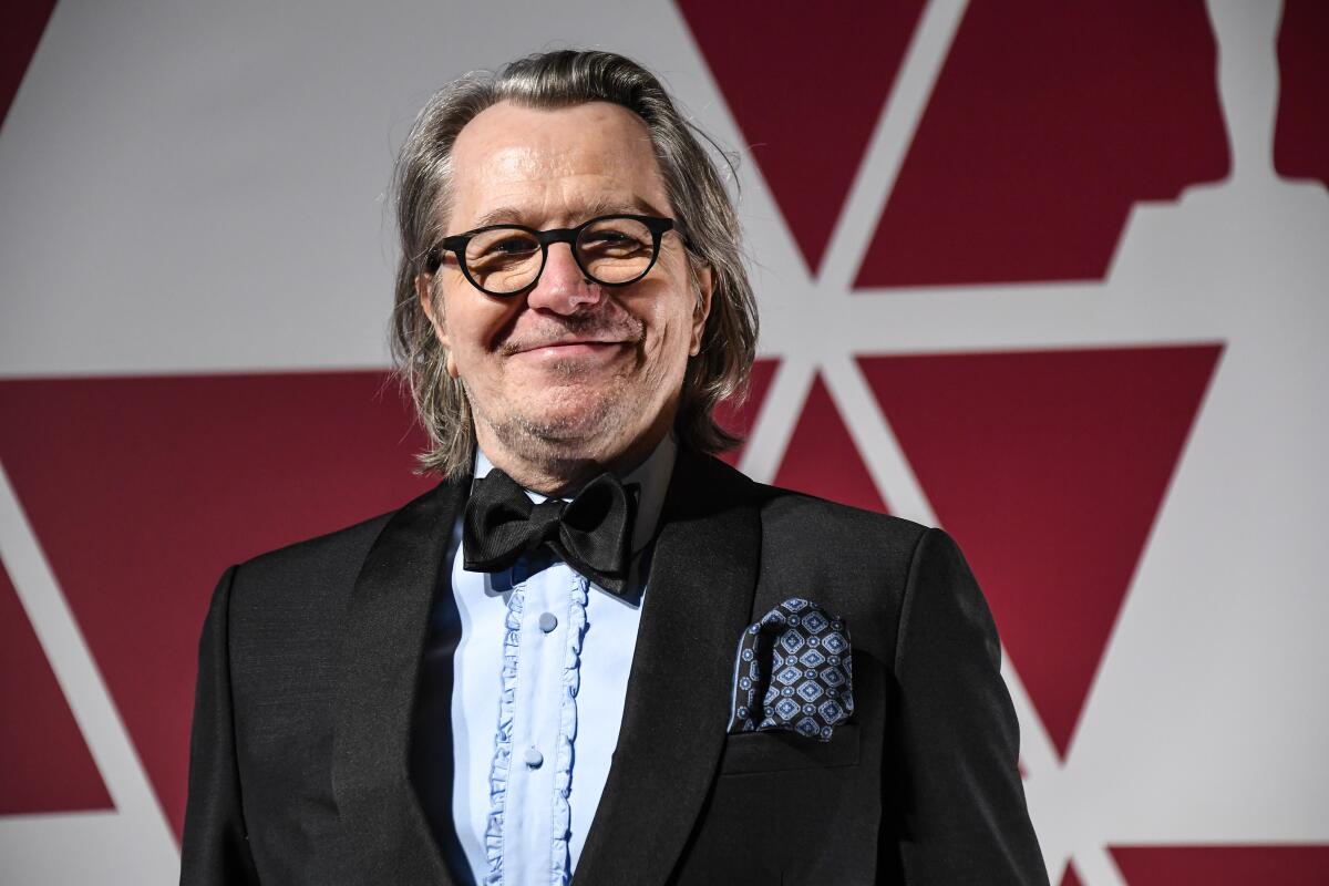 Gary Oldman smiles while wearing a black tuxedo over a light-blue shirt with ruffles