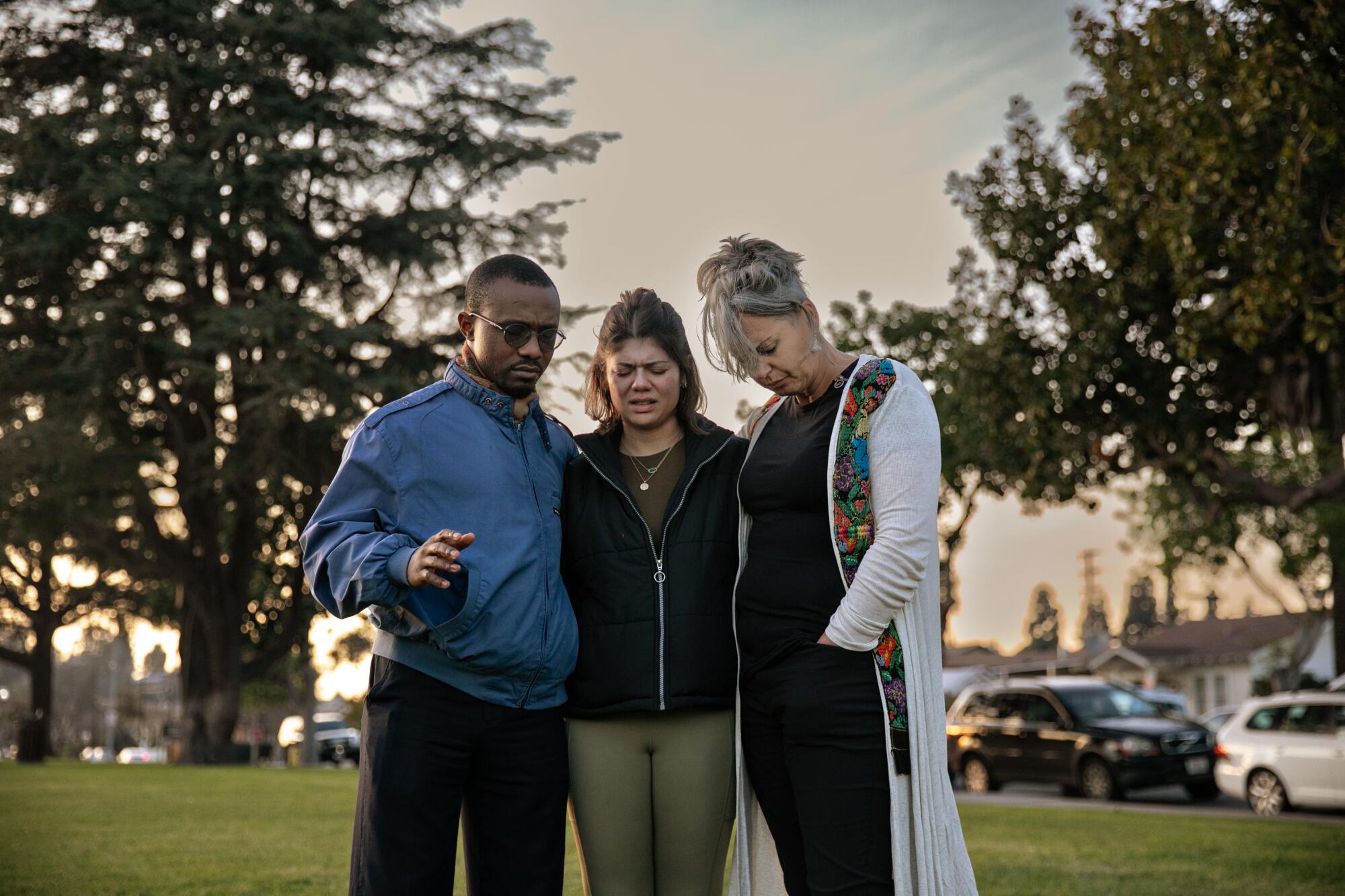 Three people stand together and pray in a grassy area.