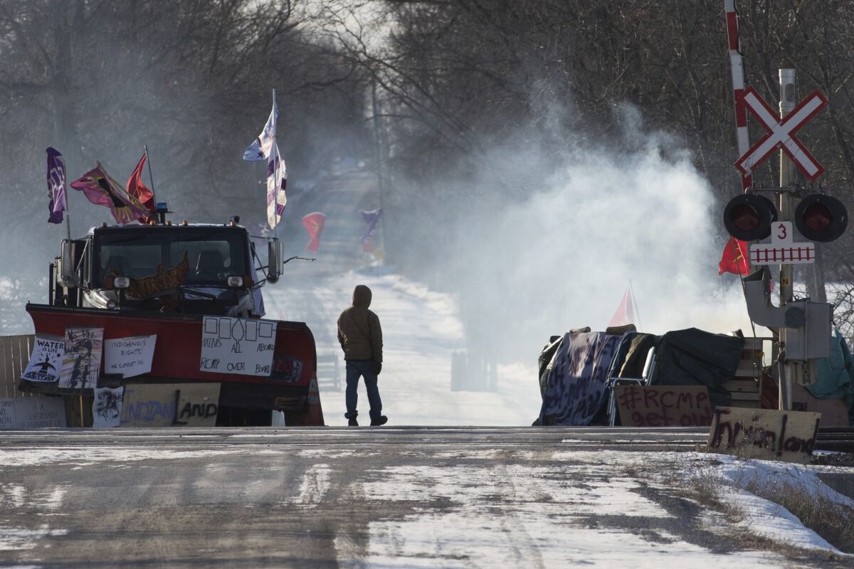 A protester stands at closed train tracks amid smoke, signs and a parked snowplow.