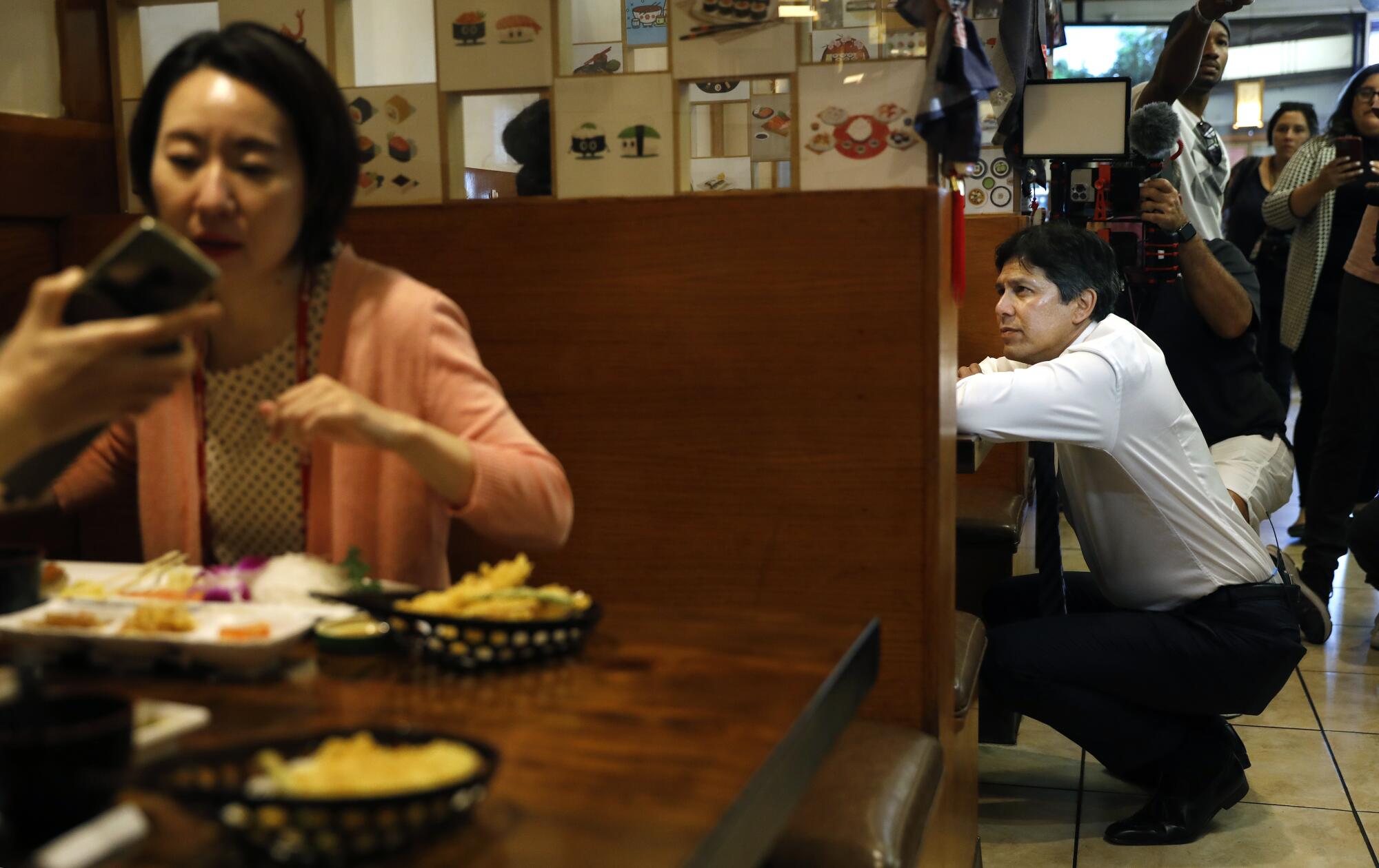 Kevin de León squatting down with his elbows on a table as he chats with patrons in a restaurant booth