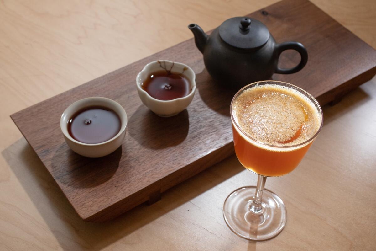 A teapot and two cups of tea rest on a wooden plant next to a cocktail in a glass.