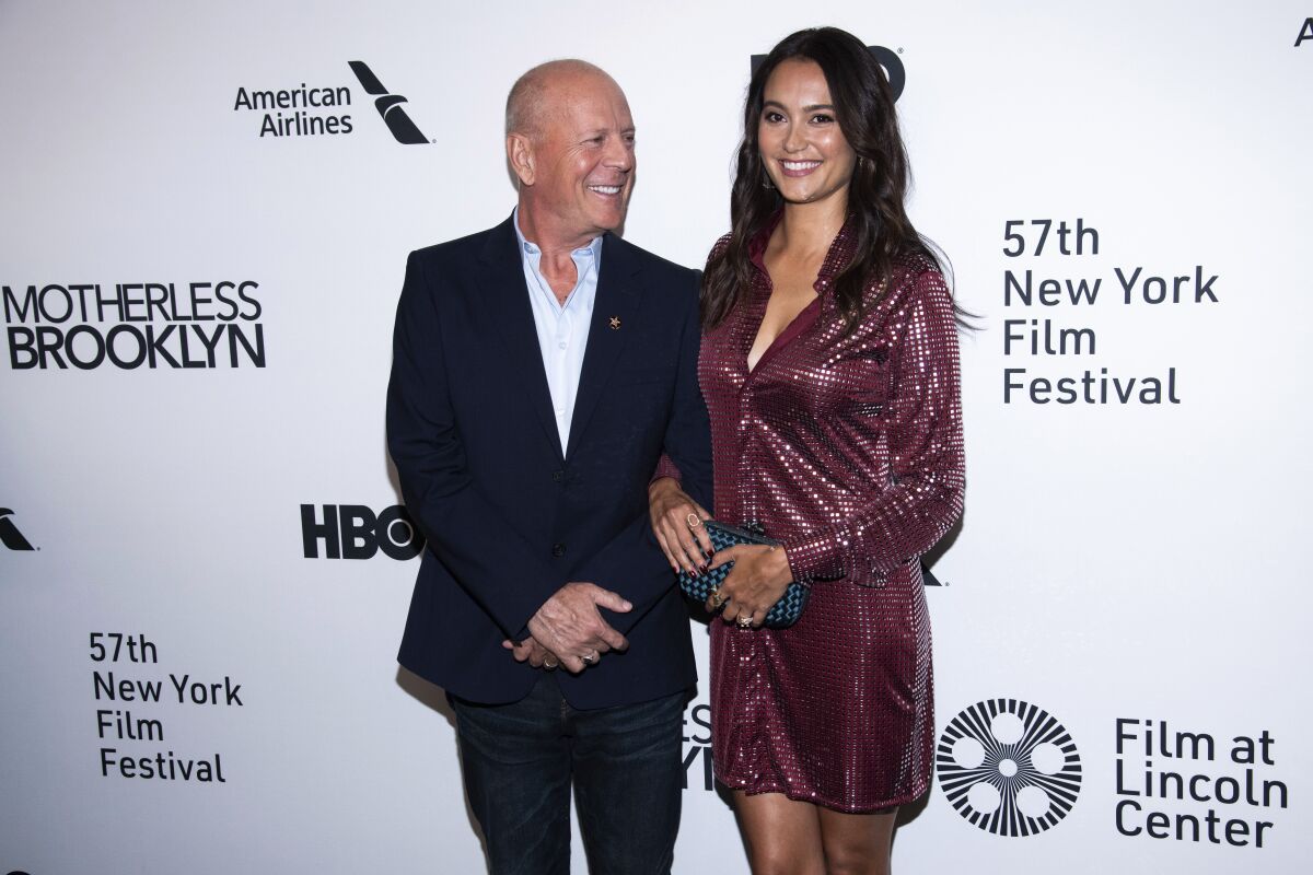 A smiling man stands with a woman, their arms linked, on a red carpet.