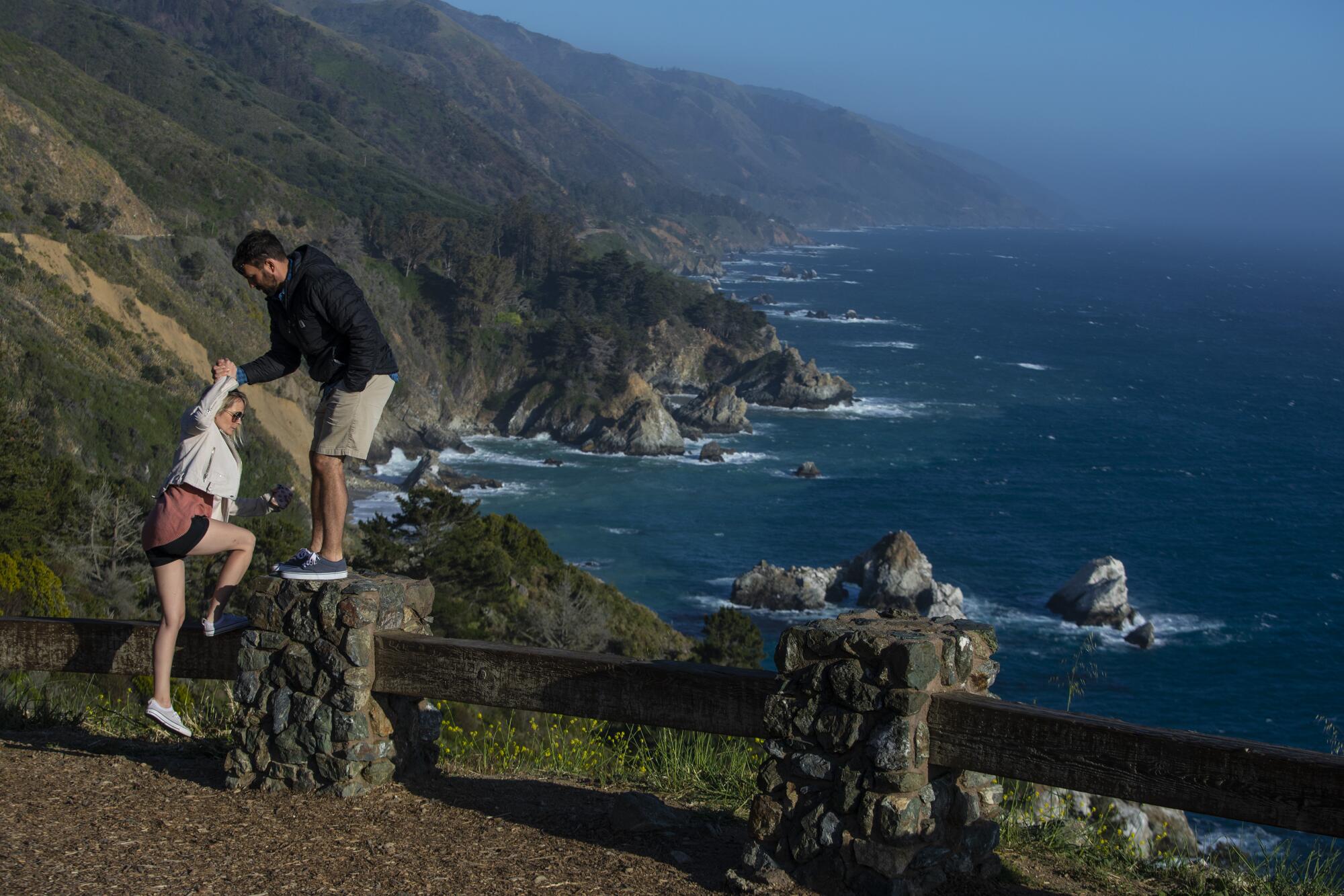 A man helps a woman climb atop a low fence at a viewing spot, with ocean and rugged coast in the background.
