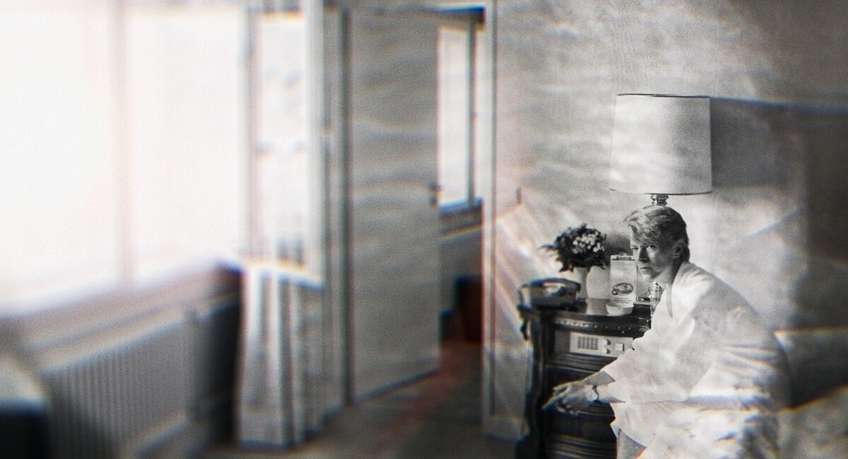 A black-and-white image of David Bowie sitting in a bedroom.