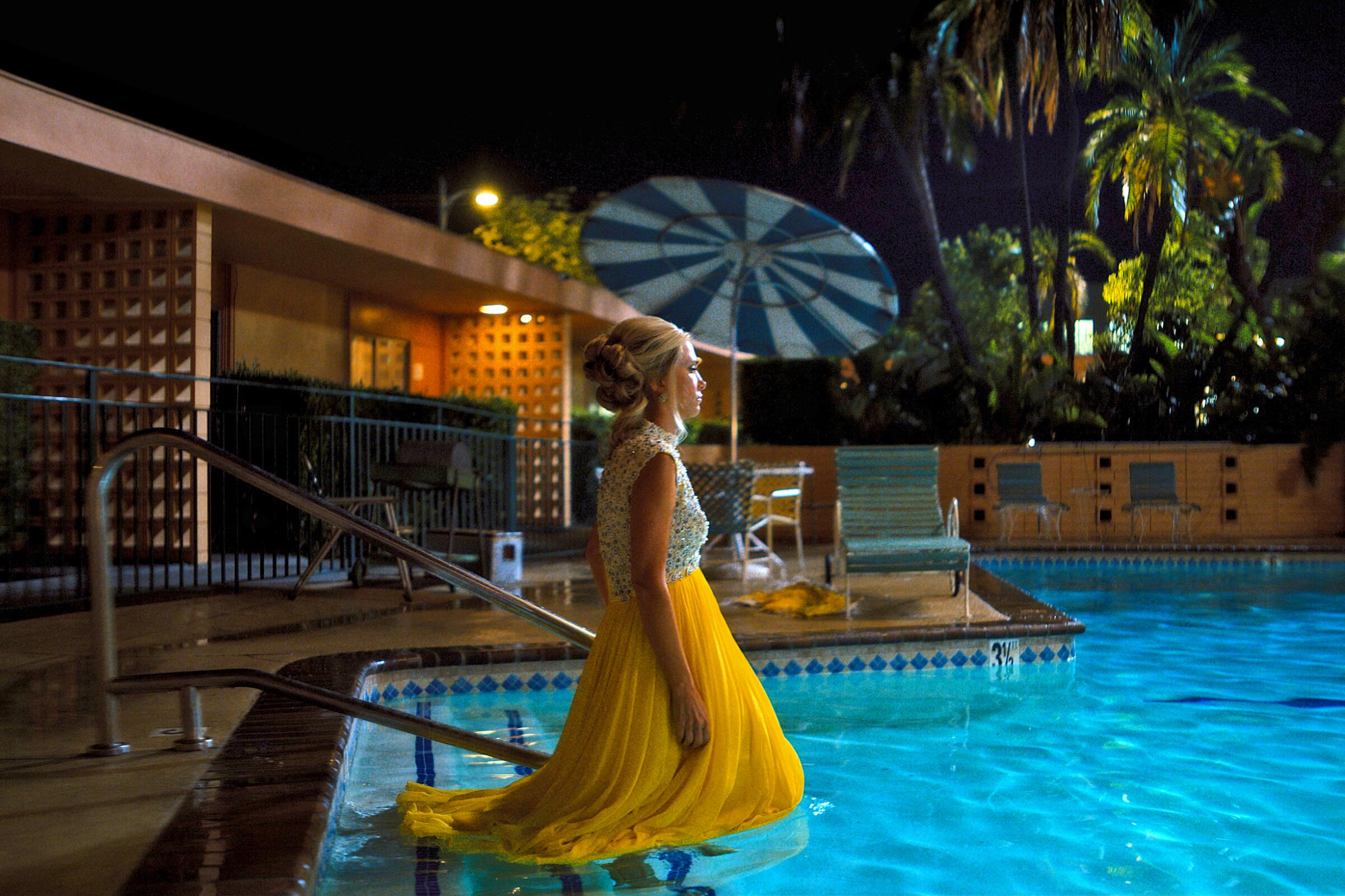 A woman in a yellow dress walks into a swimming pool at night