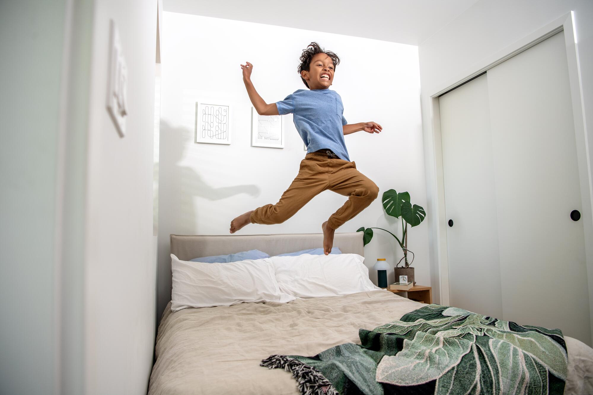 A 7-year-old boy jumps on a bed in a narrow, white-walled room