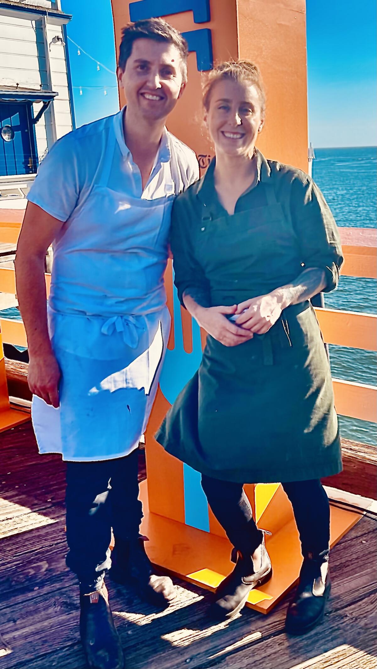 Chefs Josh Niland and Jo Barrett smiling in aprons standing on a pier.