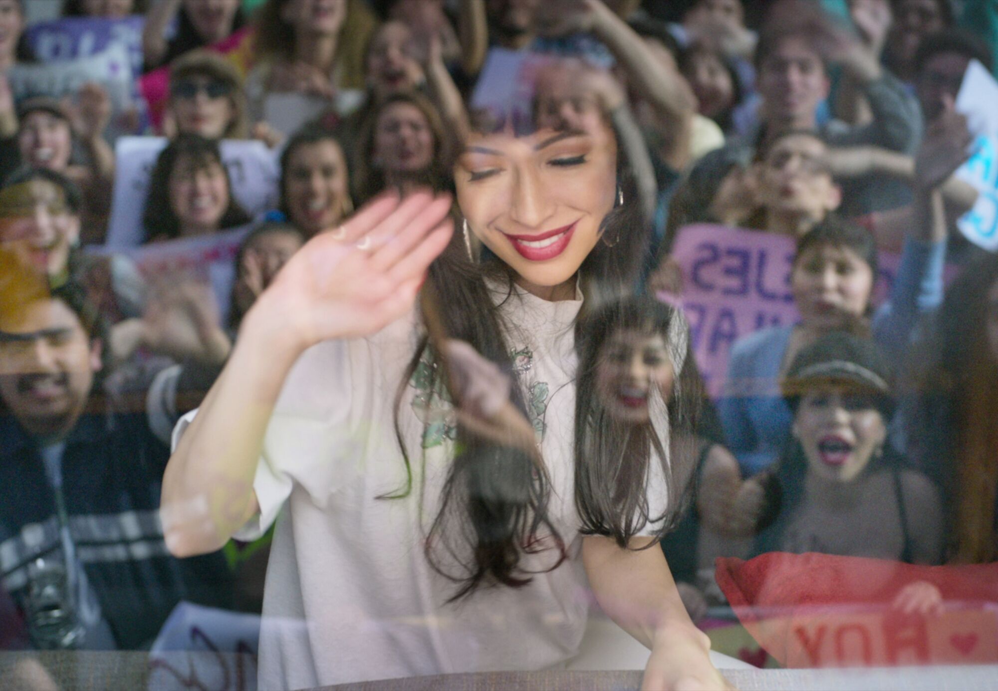 Christian Serratos, as Selena, waves to fans, who are reflected in a window.