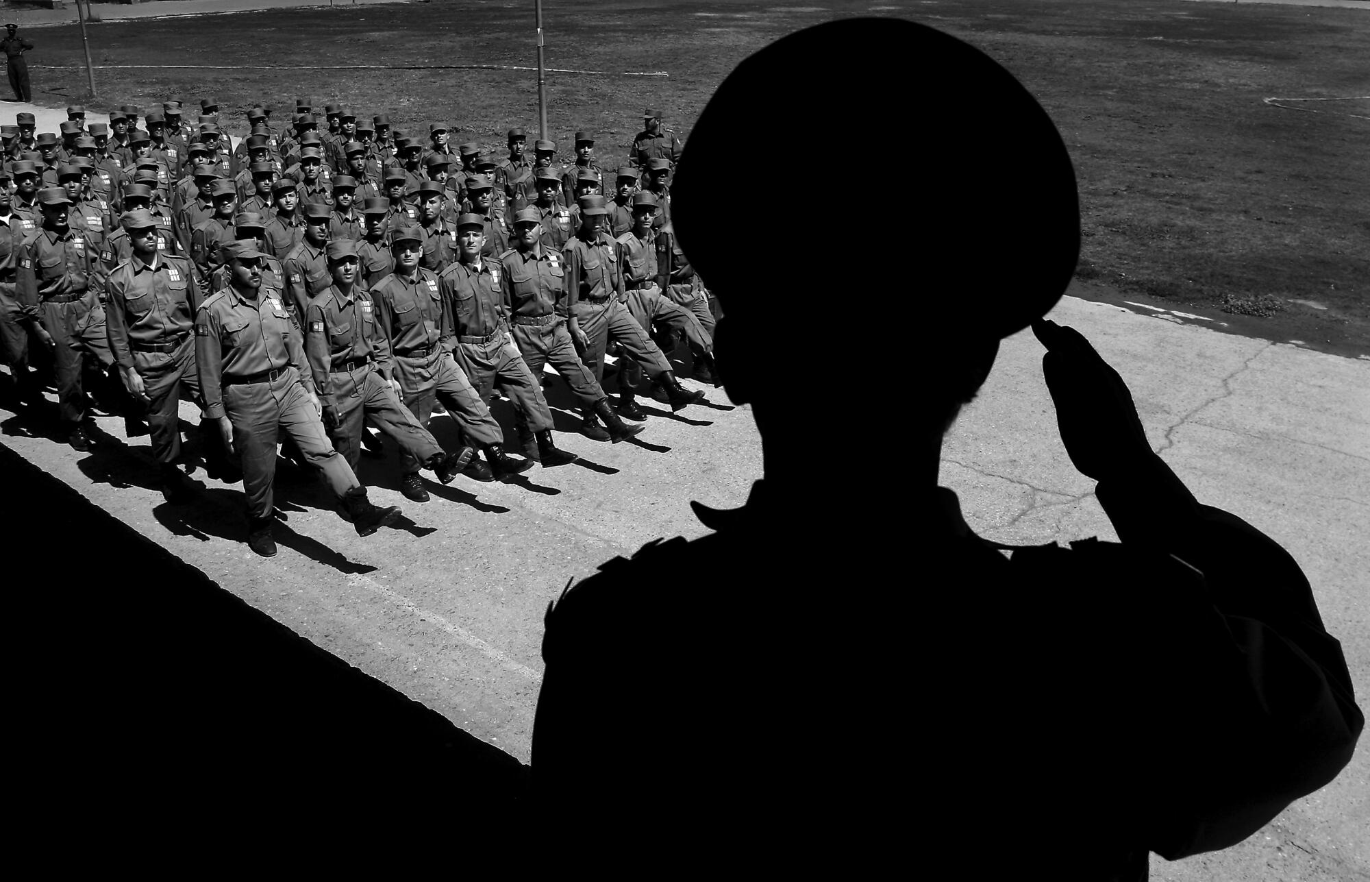 Men march in formation in front of an officer saluting them