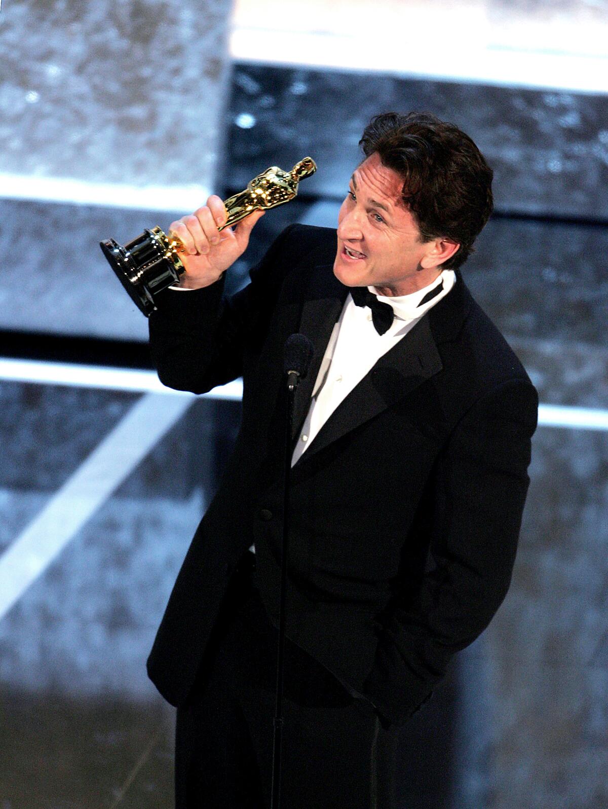A man in a tuxedo looks upward and raises his right hand, which holds an Oscar statuette