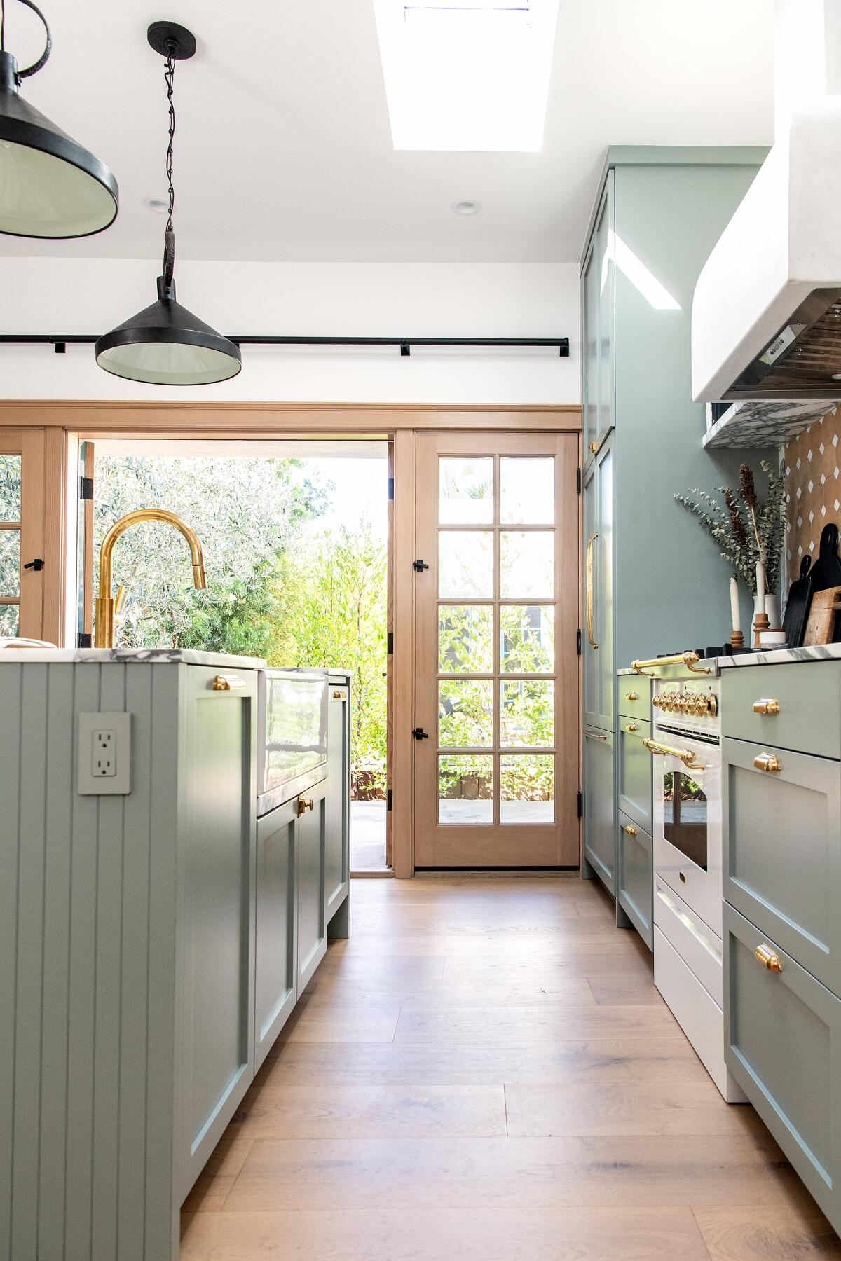 Looking through a kitchen with green-gray cabinets, gold hardware and a white oven toward an ADU's backyard.
