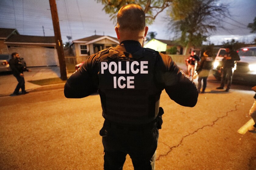 A man wearing a uniform with the words "POLICE ICE" on the back stands in a street.
