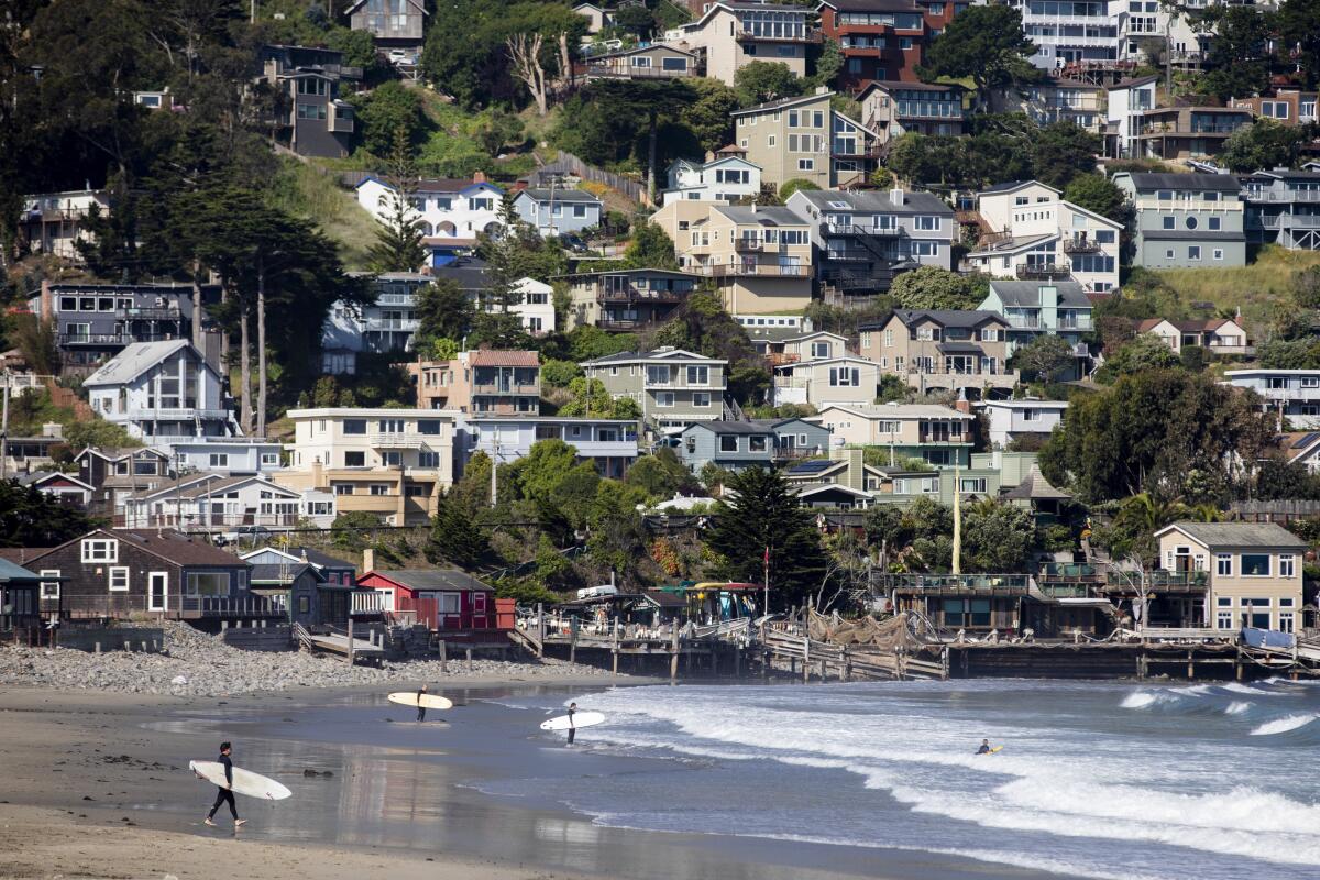Surfers walk toward the surf at the foreground, near bottom of the frame. Houses crowd the hill filling the background. 