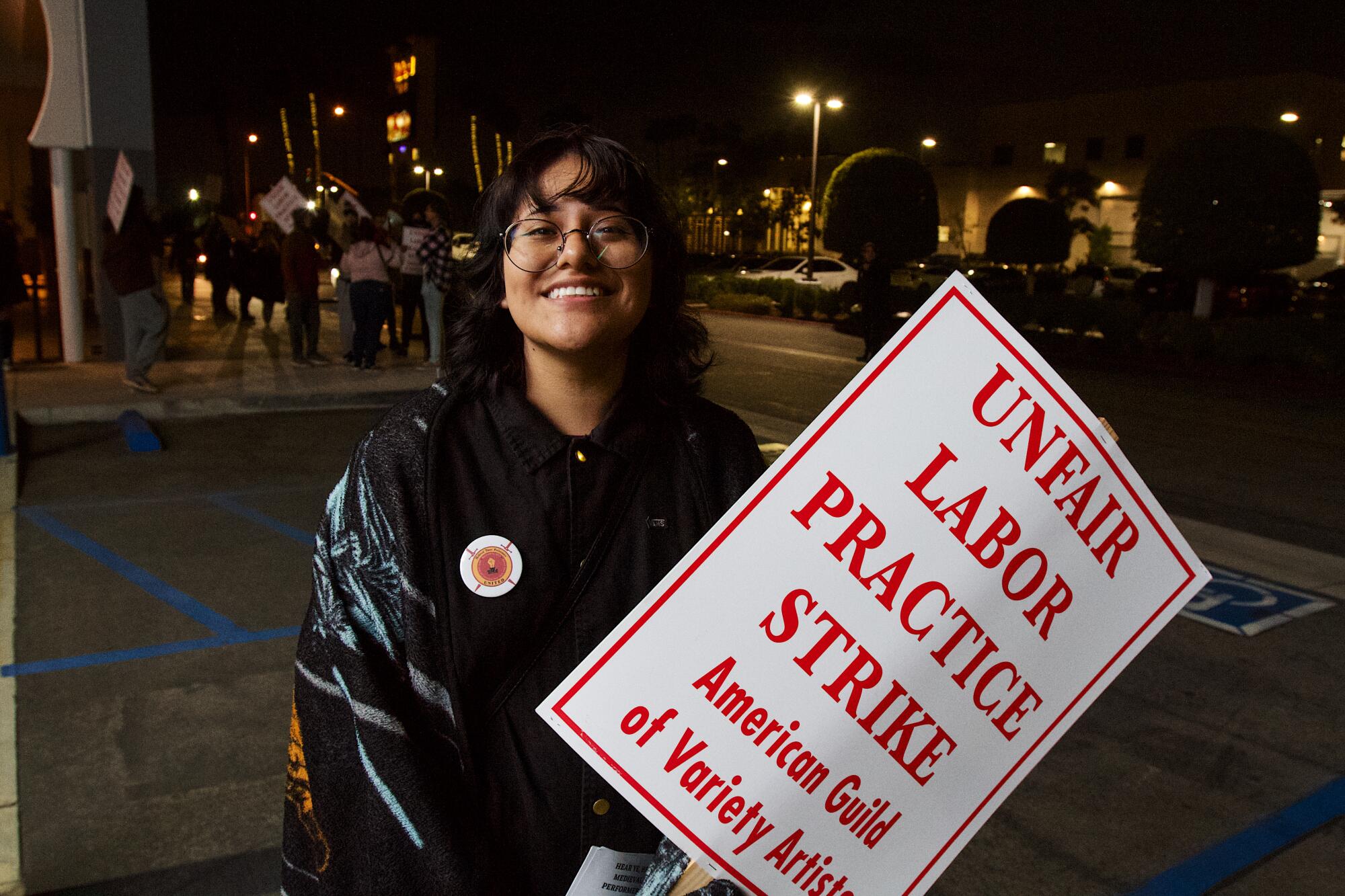 Trumpeter Lizbeth Figueroa stands smiling just outside the entrance, holding an "UNFAIR LABOR PRACTICE STRIKE" picket sign.