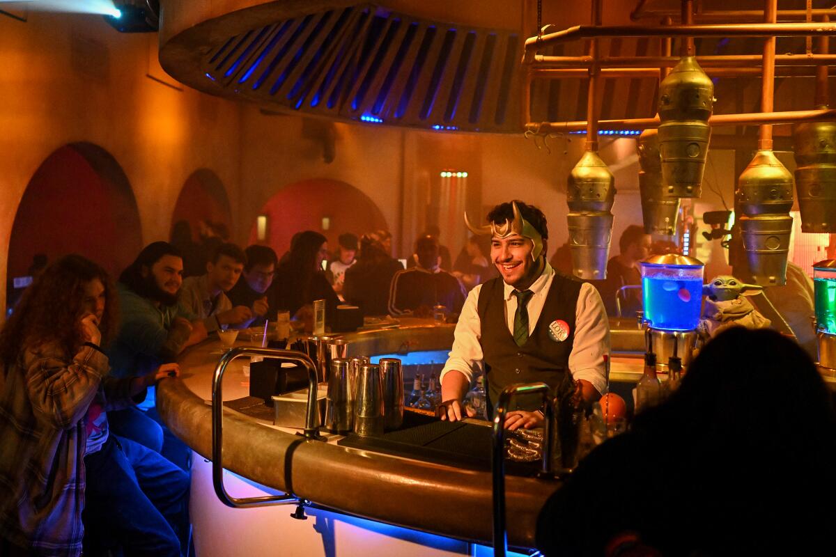 A bartender wears a horned headpiece as he stands behind a stylish sci-fi themed bar.