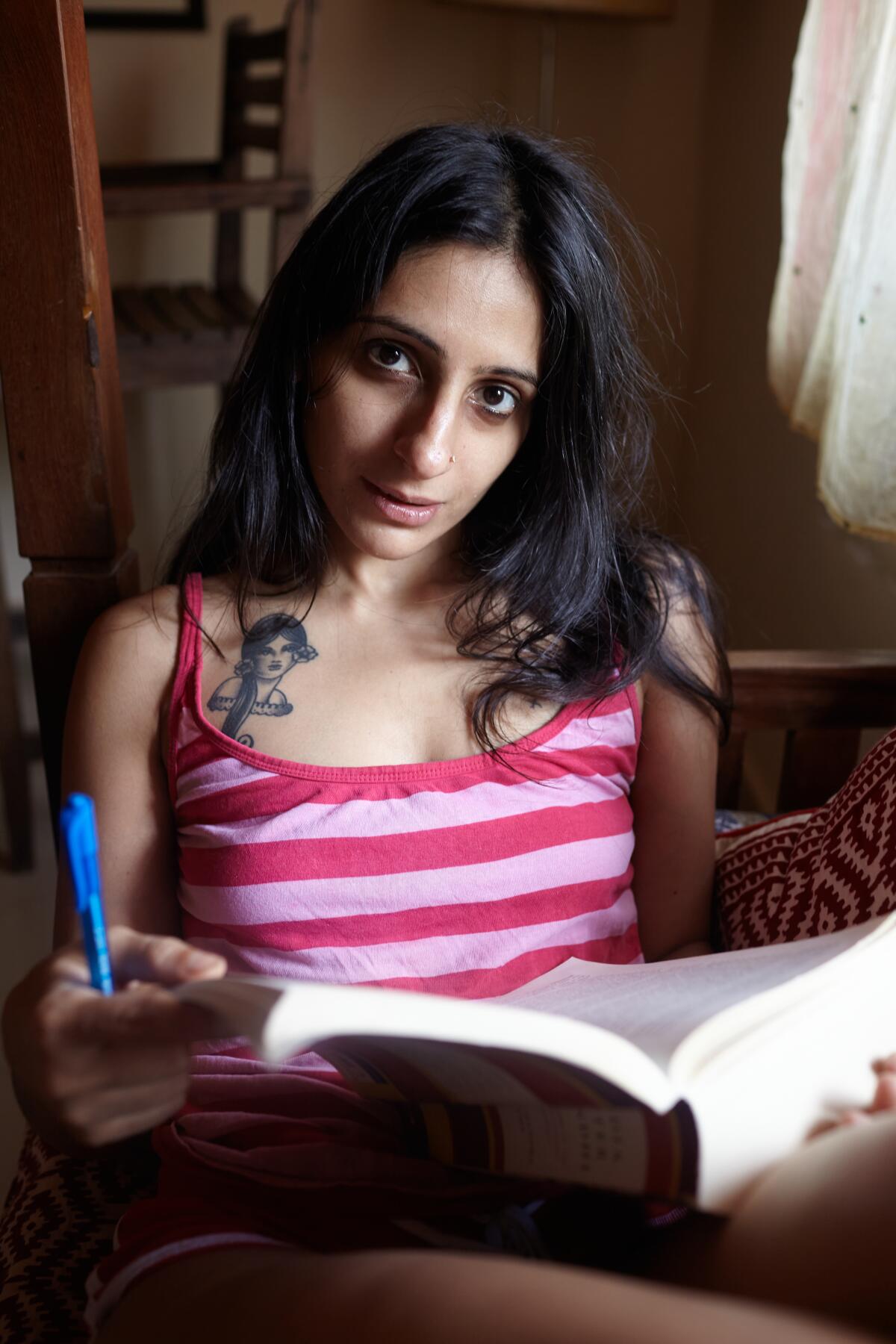 A tattooed woman in a pink and white striped tank top holds a pen and book.