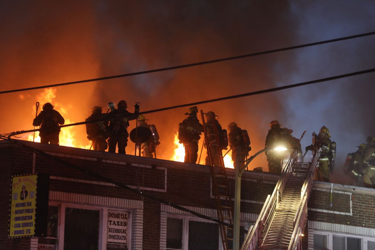 Firefighters stand on the roof of an apartment building as flames and smoke pour out of it