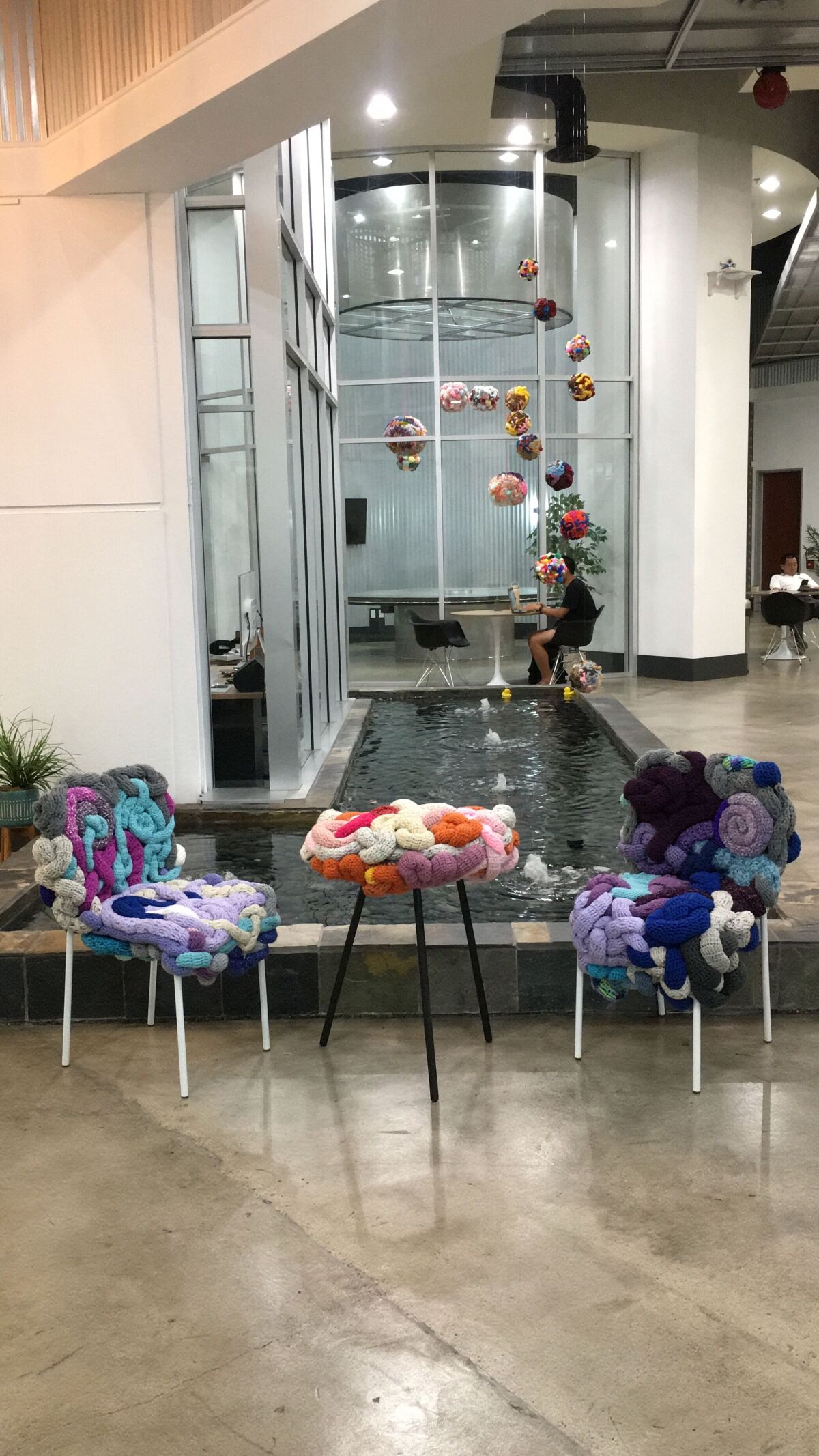 Threadwinners' "zer | o" exhibit, presented by Aquila Projects at Irvine's WorkWell, decorates multiple rooms of the co-working space, including a set of tube chairs and a side table. Behind are hanging yarn sculptures over the office's indoor fountain.