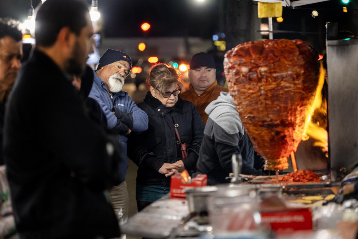 Customers wait for tacos as meat cooks on a vertical rotisserie.
