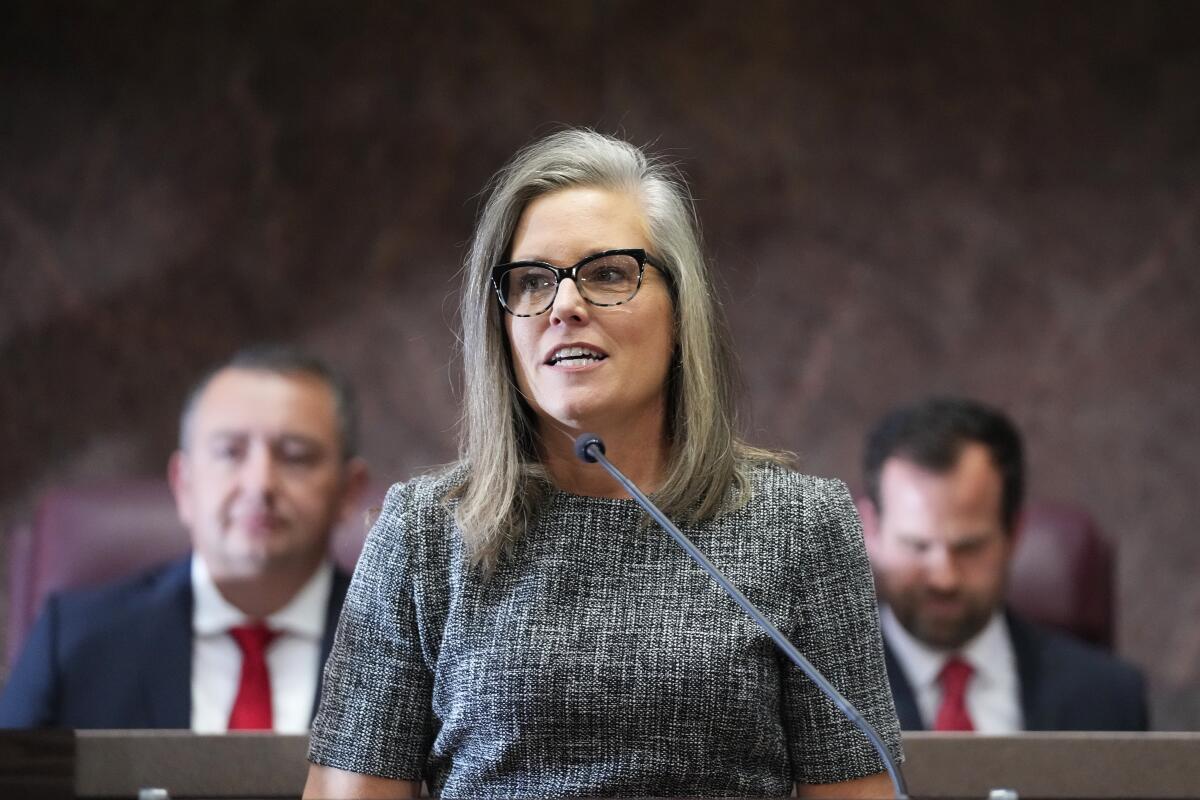 Arizona Gov. Katie Hobbs standing and speaking into a microphone, two men seated behind her