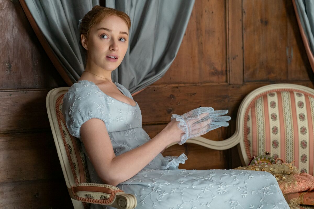 A young woman sitting in an eggshell blue Empire dress, removing a lace glove