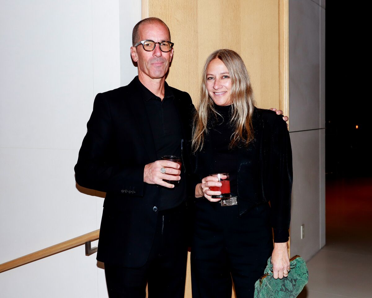 A man and a woman, dressed in black, stand holding drink glasses and smiling for the camera.