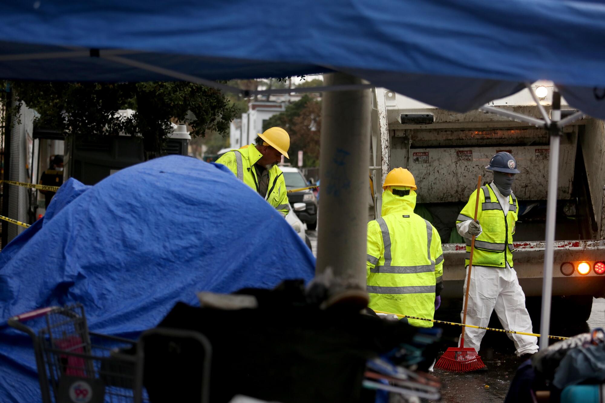 Workers in hard hats and reflective jackets stand near a trash truck and items covered by blue tarps 