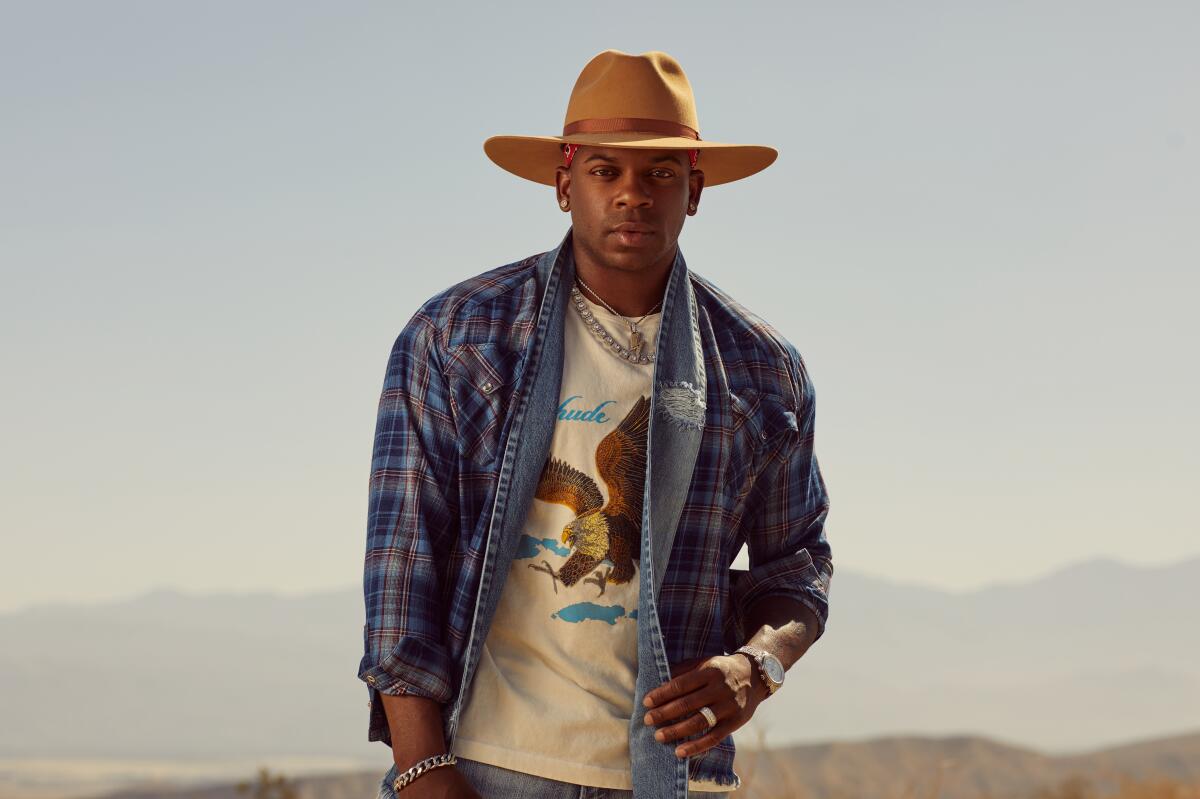 Country musician Jimmie Allen stands outdoors in a cowboy hat and a denim-and-flannel jacket