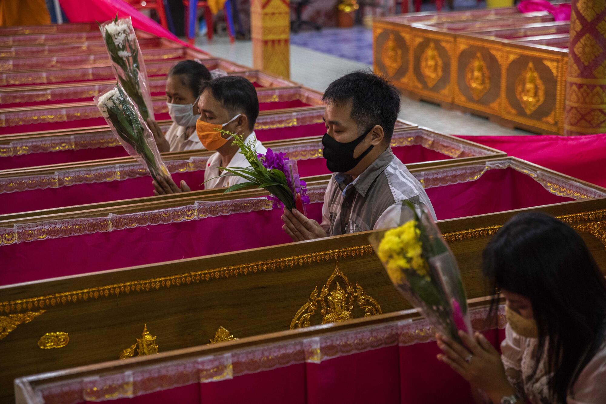 People in masks sit inside coffins, holding bouquets of flowers and praying.
