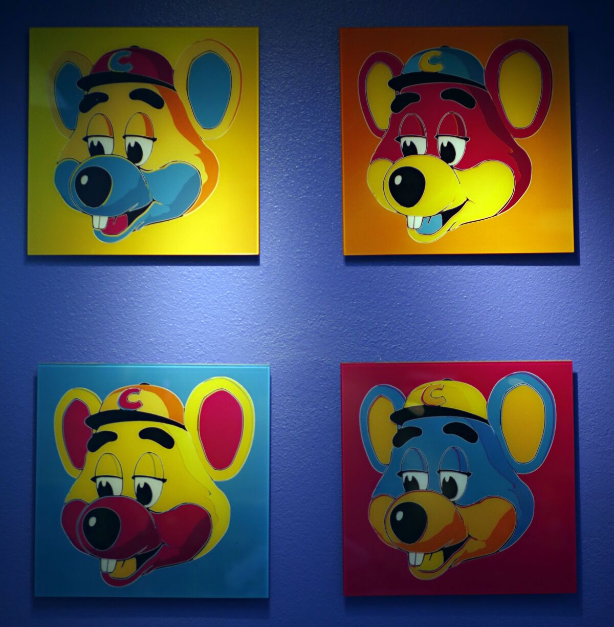Four colorful paintings of Chuck E. Cheese hang on a blue wall.