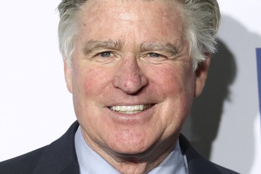 Treat Williams with gray combed hair is smiling while wearing a black suit with a light blue shirt and gray tie. 