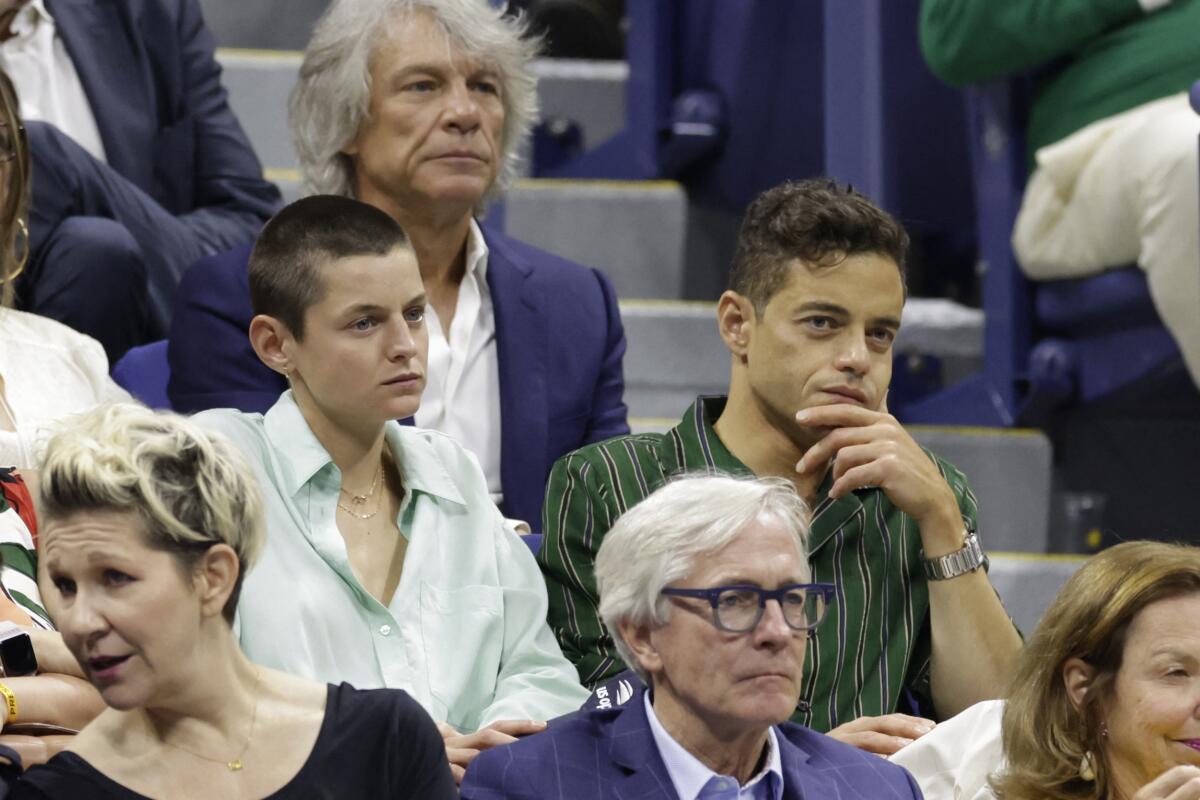 Emma Corrin, in a button-down shirt with buzzed hair, sits with Rami Malek, in a green striped shirt with a hand on his chin