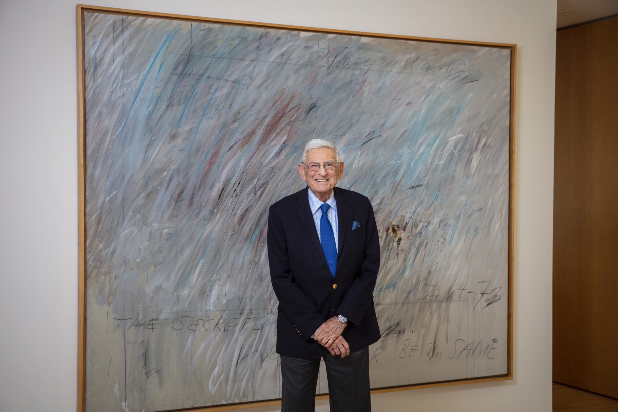 Eli Broad, in a suit and tie, stands in front of a large abstract painting. 