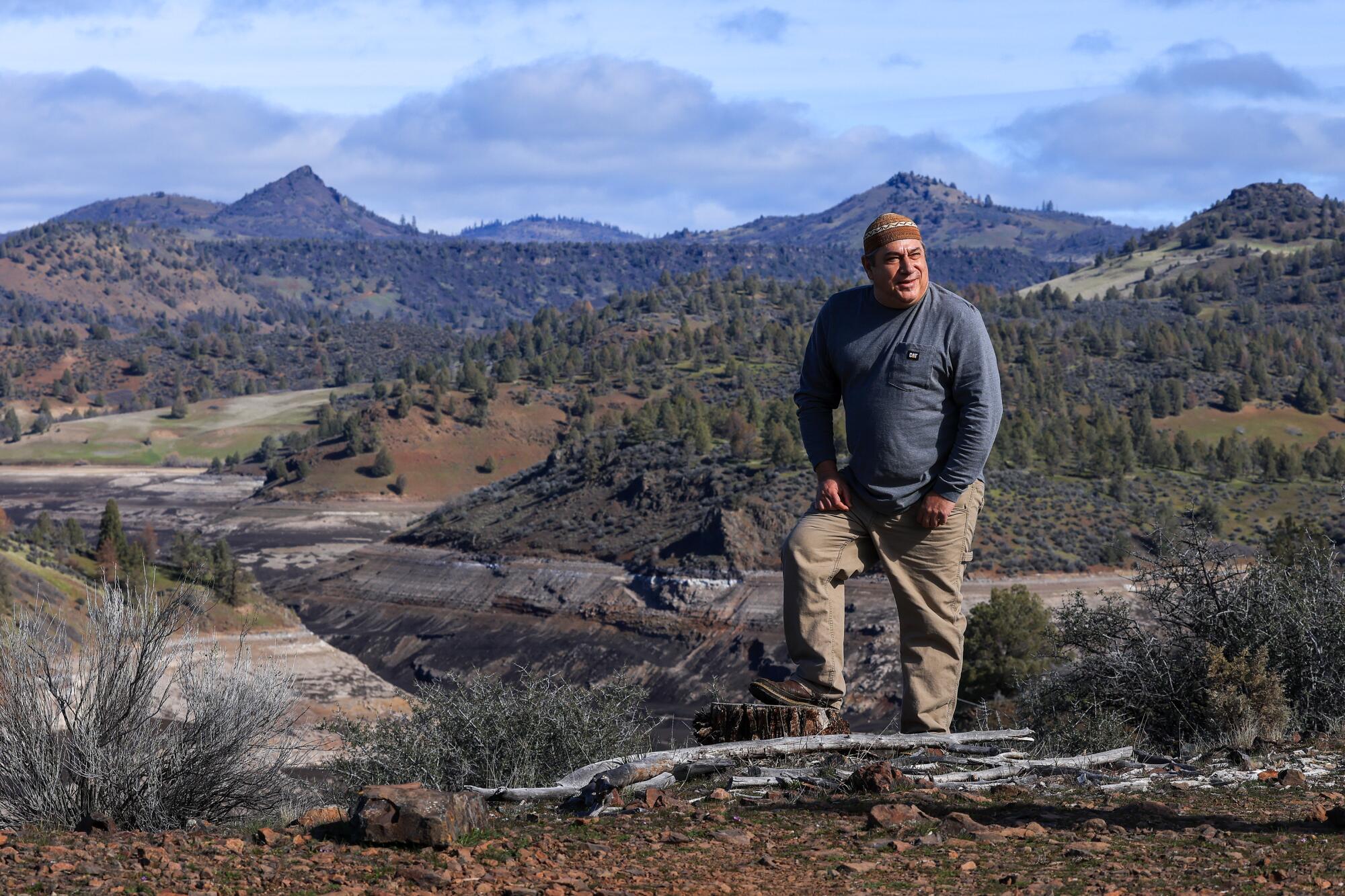 A man standing on a bluff, with a muddy reservoir, evergreen trees and hills behind him