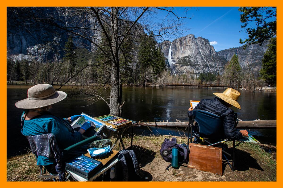At the waters edge, two people wearing hats  paint a scene of Yosemite Falls