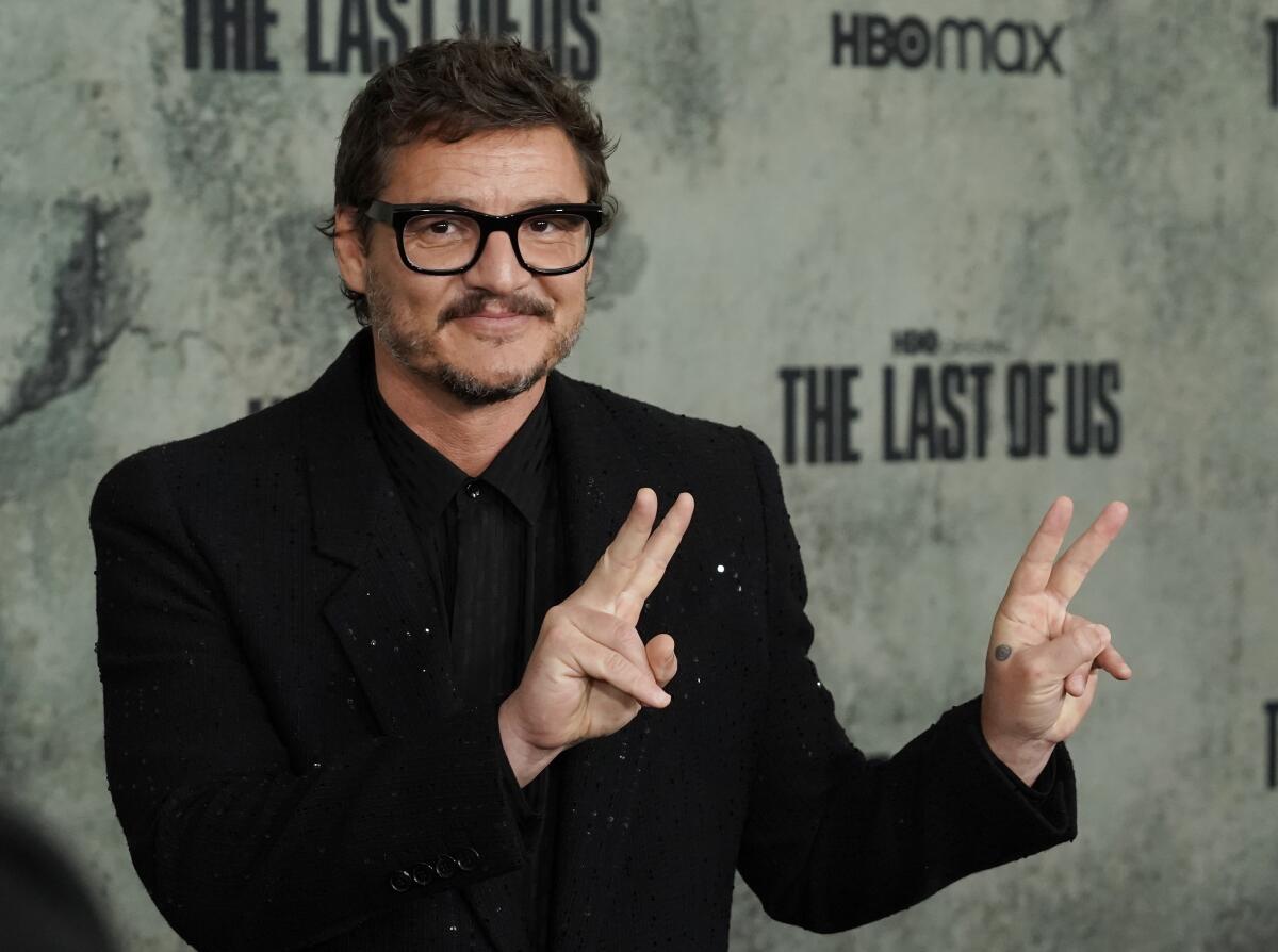 A man wearing black glasses and a black suit holds up peace signs with both hands