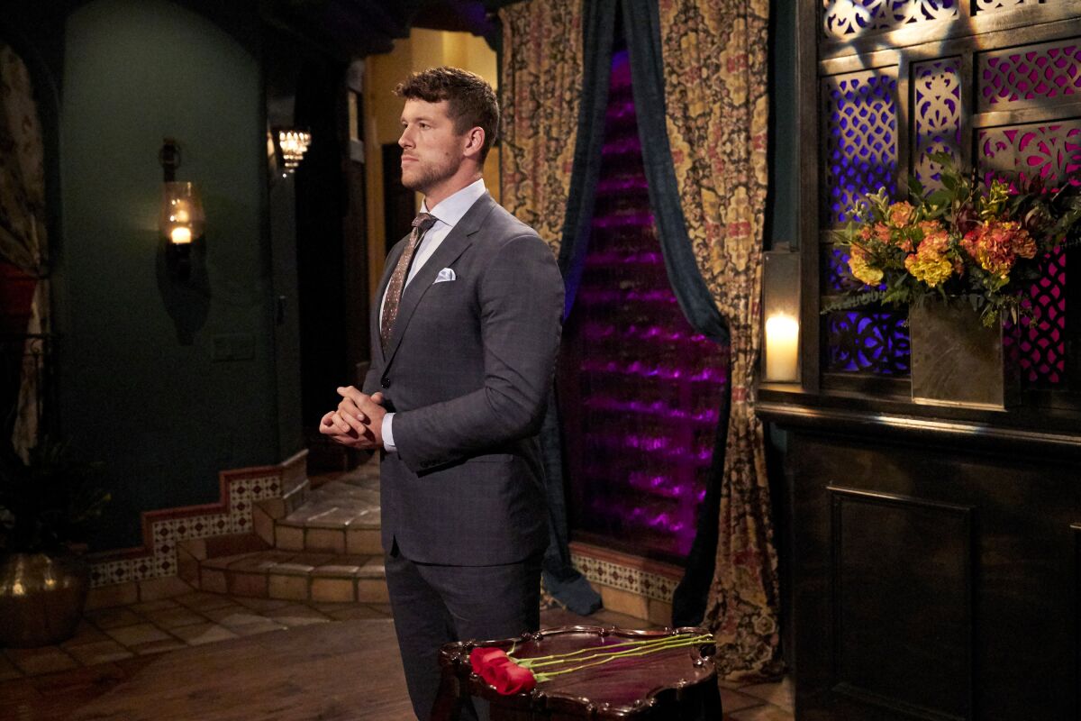 A man in a suit, looking serious, stands in an opulently decorated foyer.