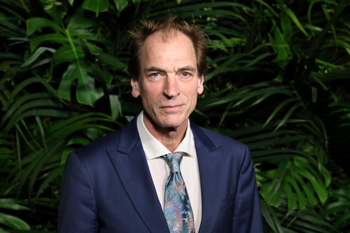 Julian Sands in a head-and-shoulders frame looks at the camera while standing in front of greenery