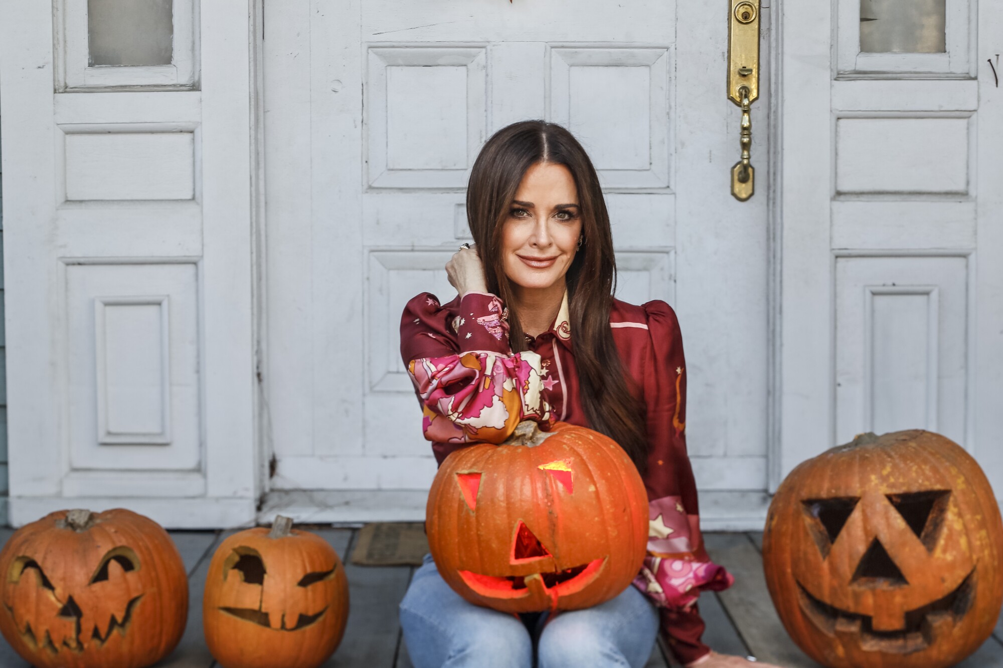 A woman sits on the front steps of a home flanked by jack-o'-lanterns with another one on her lap