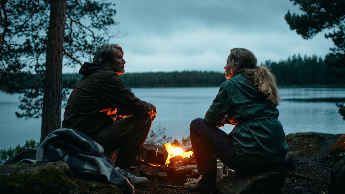 A husband and wife sit by a campfire beside a lake.